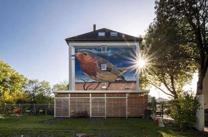 Das Wandbild von Denis Klatt zeigt ein überdimensionales Rotkehlchen, das in einer surrealen Szenerie aus Wüste und Meer eingebettet ist. Das Wandbild von Denis Klatt zeigt ein überdimensionales Rotkehlchen, das in einer surrealen Szenerie aus Wüste und Meer eingebettet ist. (vergrößerte Bildansicht wird geöffnet)